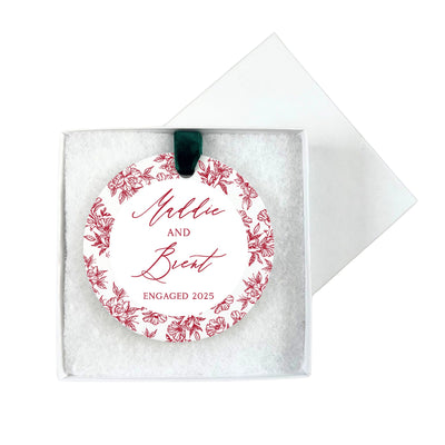 A circular white engagement ornament featuring a red floral border design, suspended by a green velvet ribbon, and presented in an open white gift box.