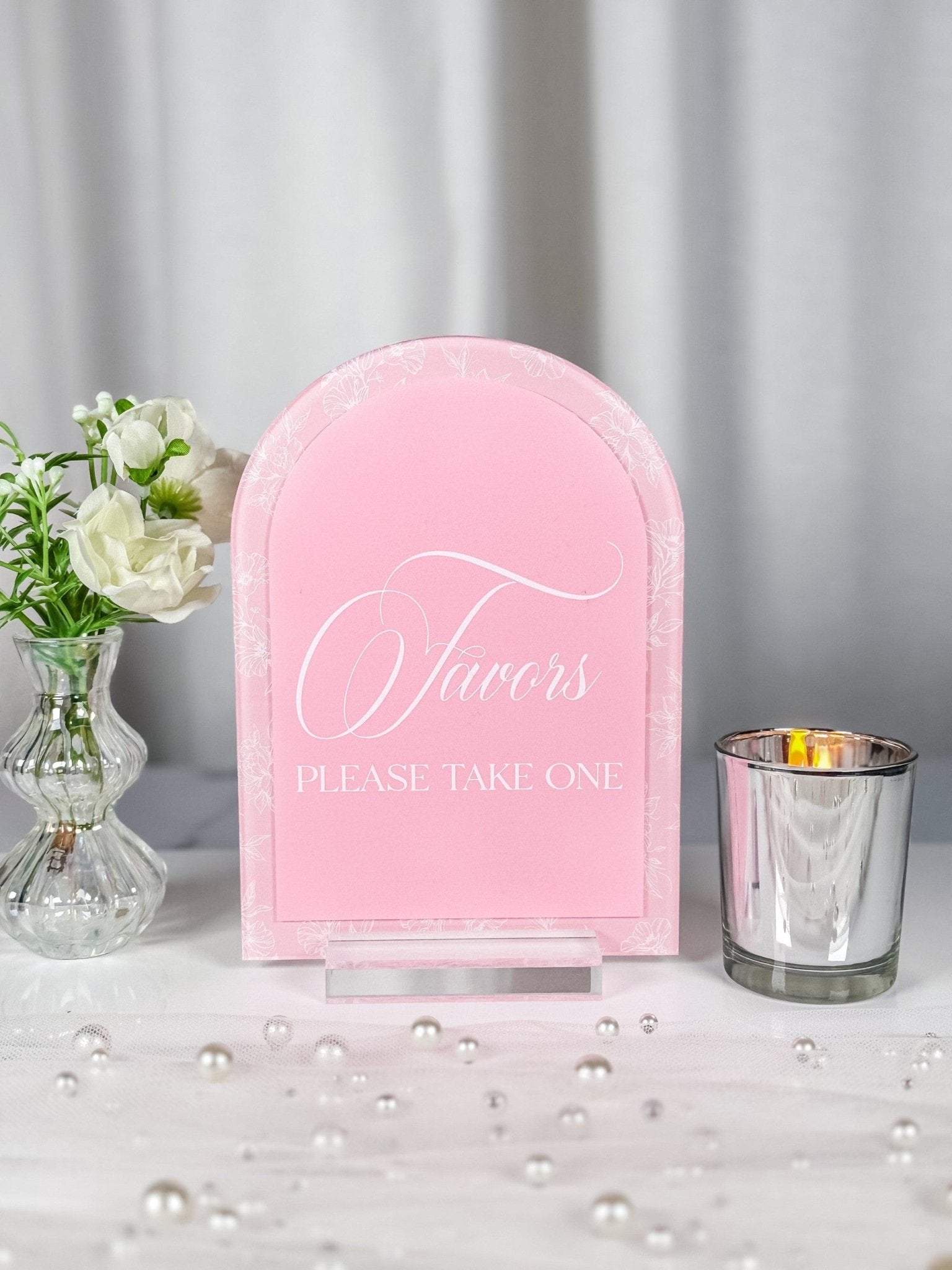 An elegant arch-shaped pink acrylic favor sign, featuring a delicate white floral border, stands on a clear base as part of event decor.