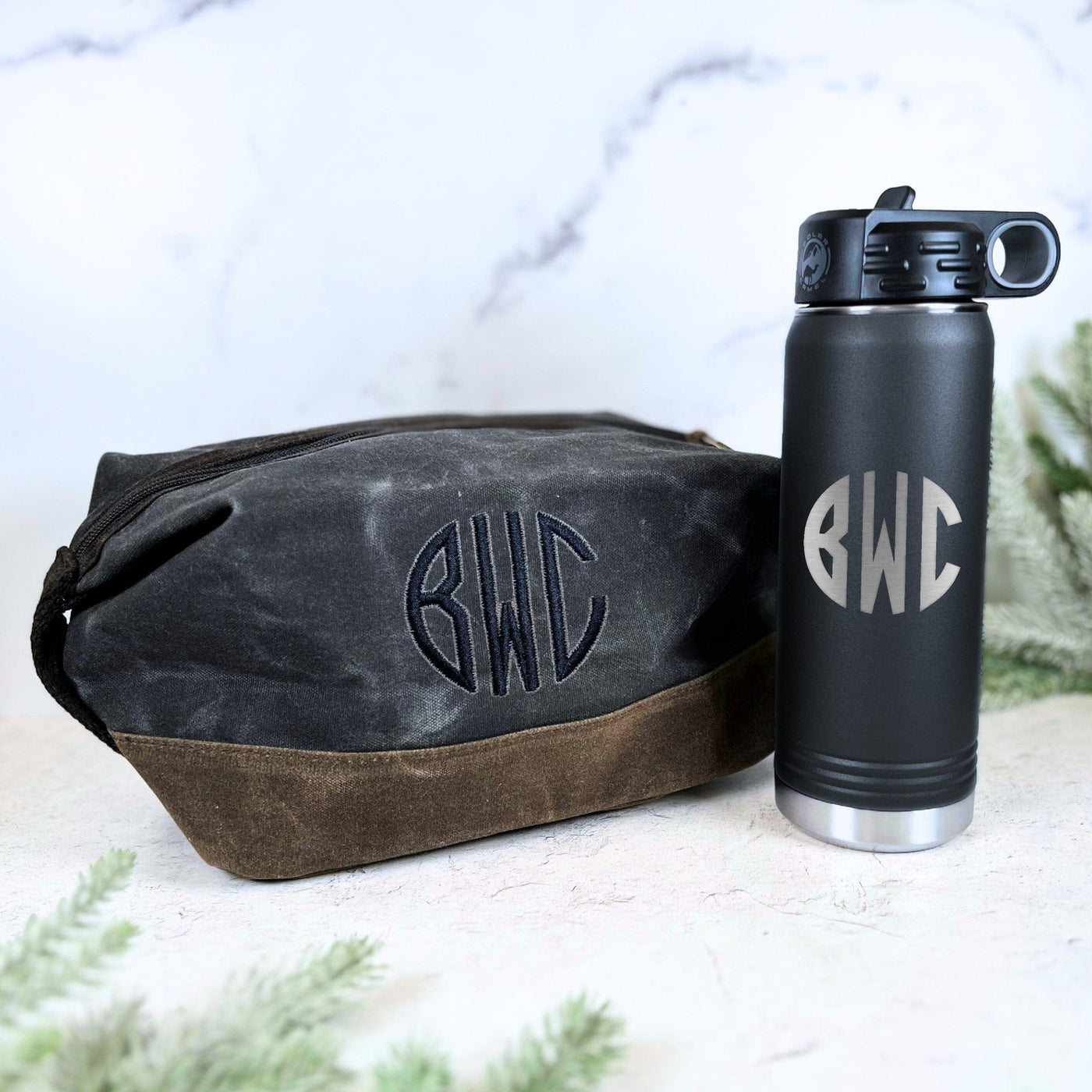 A personalized gift set featuring a black insulated stainless steel water bottle and a dark grey waxed canvas toiletry bag with a brown suede bottom, both displaying custom monograms.