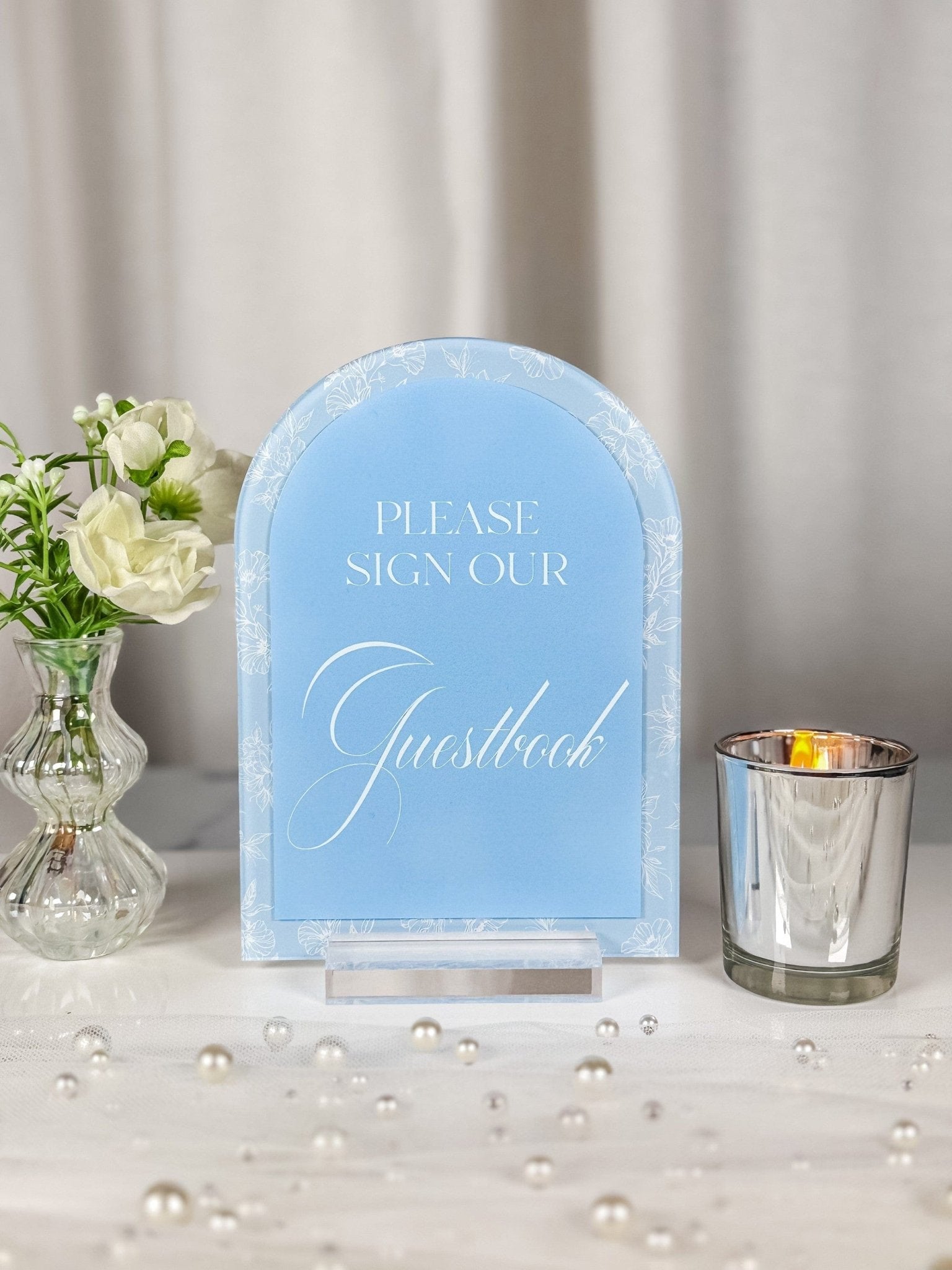 An elegant arched acrylic guestbook sign featuring a light blue background with a delicate white floral border, designed to guide guests at a wedding or special event.