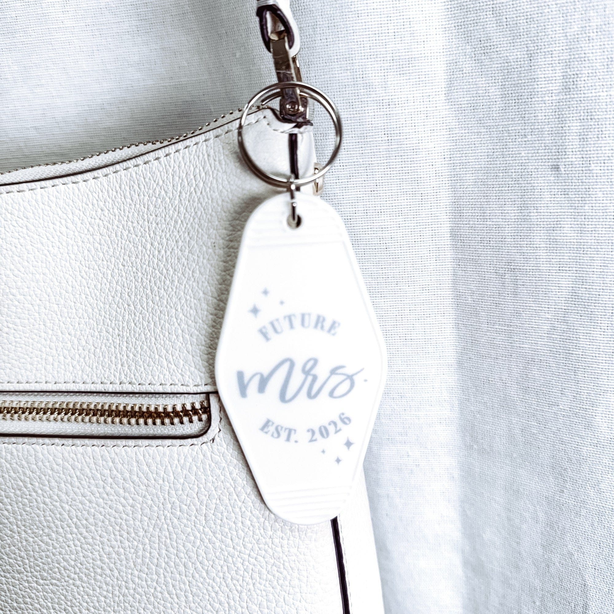 A white motel-style acrylic keytag with light gray text and sparkling accents is attached to a white leather bag, serving as a bridal accessory.
