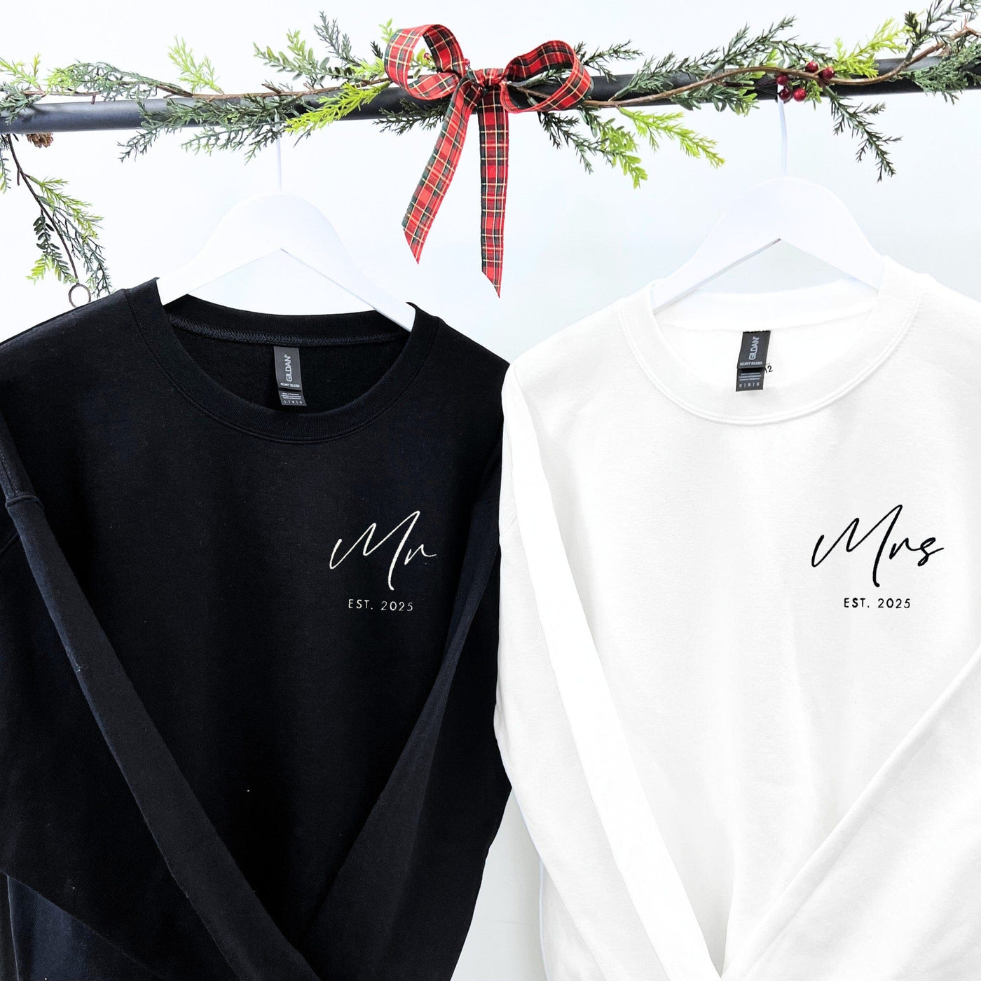 A pair of black and white long-sleeved crewneck sweatshirts, displayed on white hangers with festive greenery, designed as a personalized apparel gift for a couple.