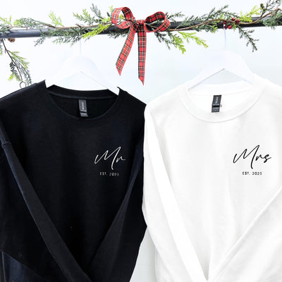 A pair of black and white long-sleeved crewneck sweatshirts, displayed on white hangers with festive greenery, designed as a personalized apparel gift for a couple.