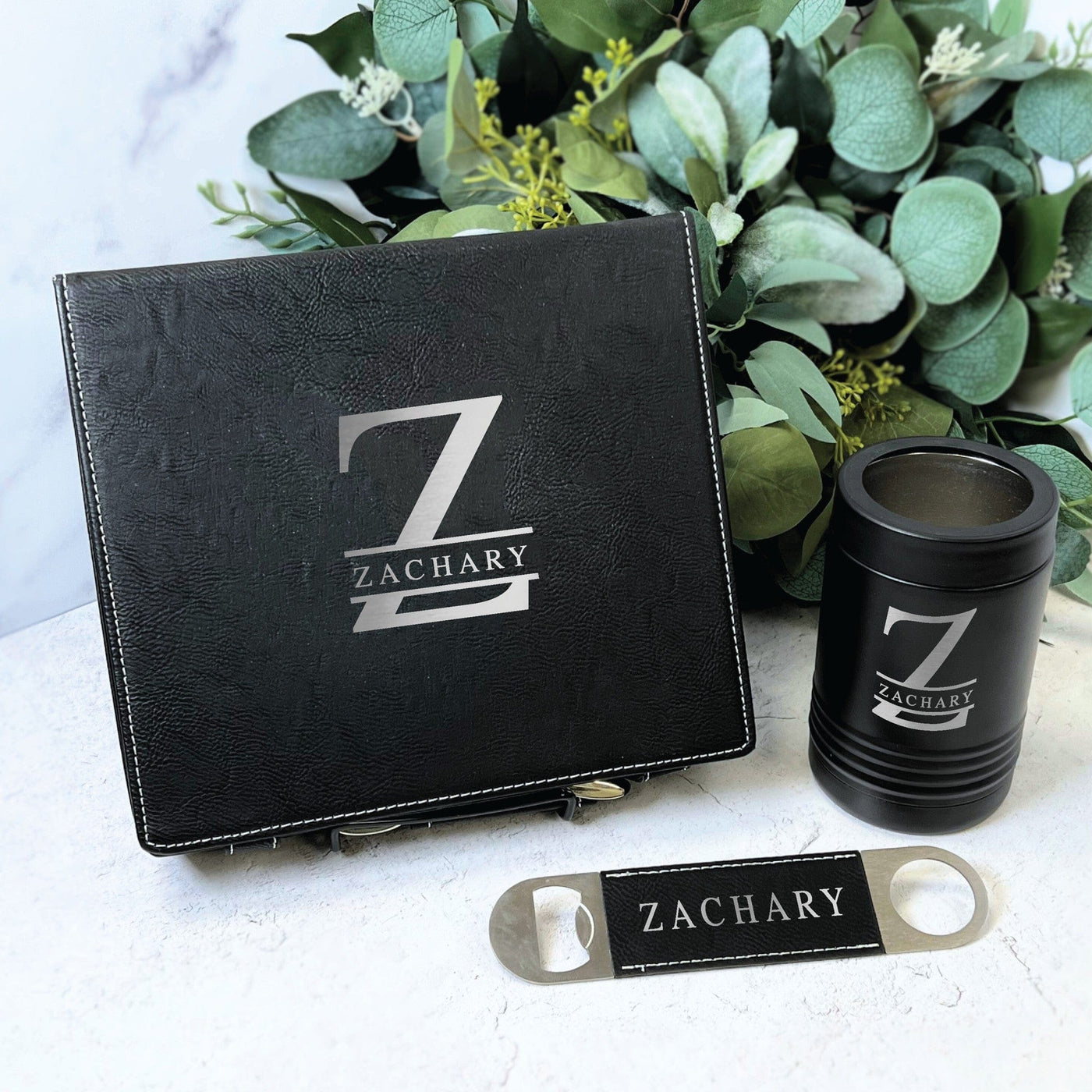 A personalized black leatherette gift box, an insulated can cooler, and a metal bottle opener, all featuring custom laser-engraved designs, ideal for a groomsmen gift or special occasion.