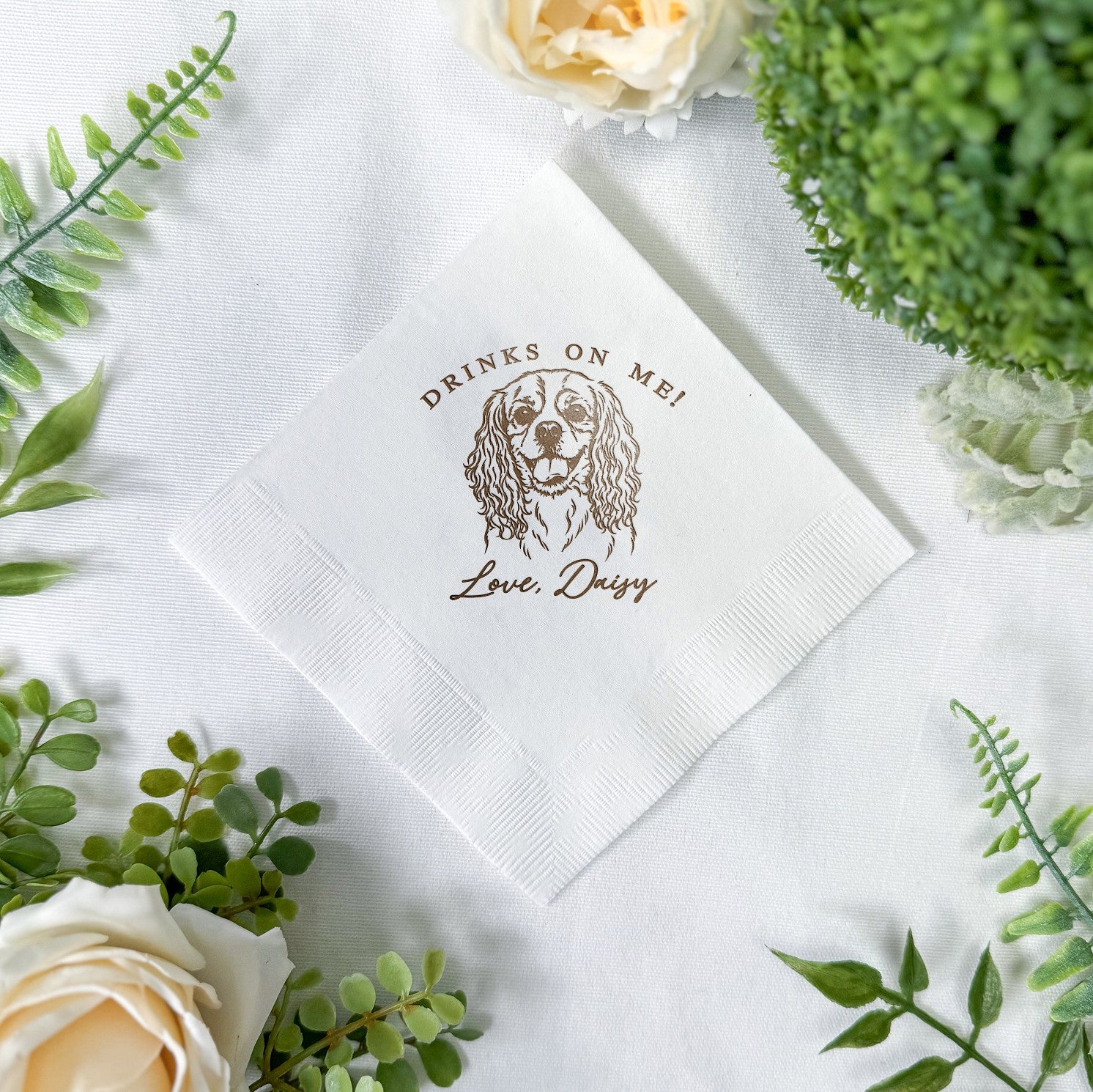 A folded white paper event napkin is decorated with a custom brown ink illustration of a dog's face, surrounded by greenery and soft cream flowers.