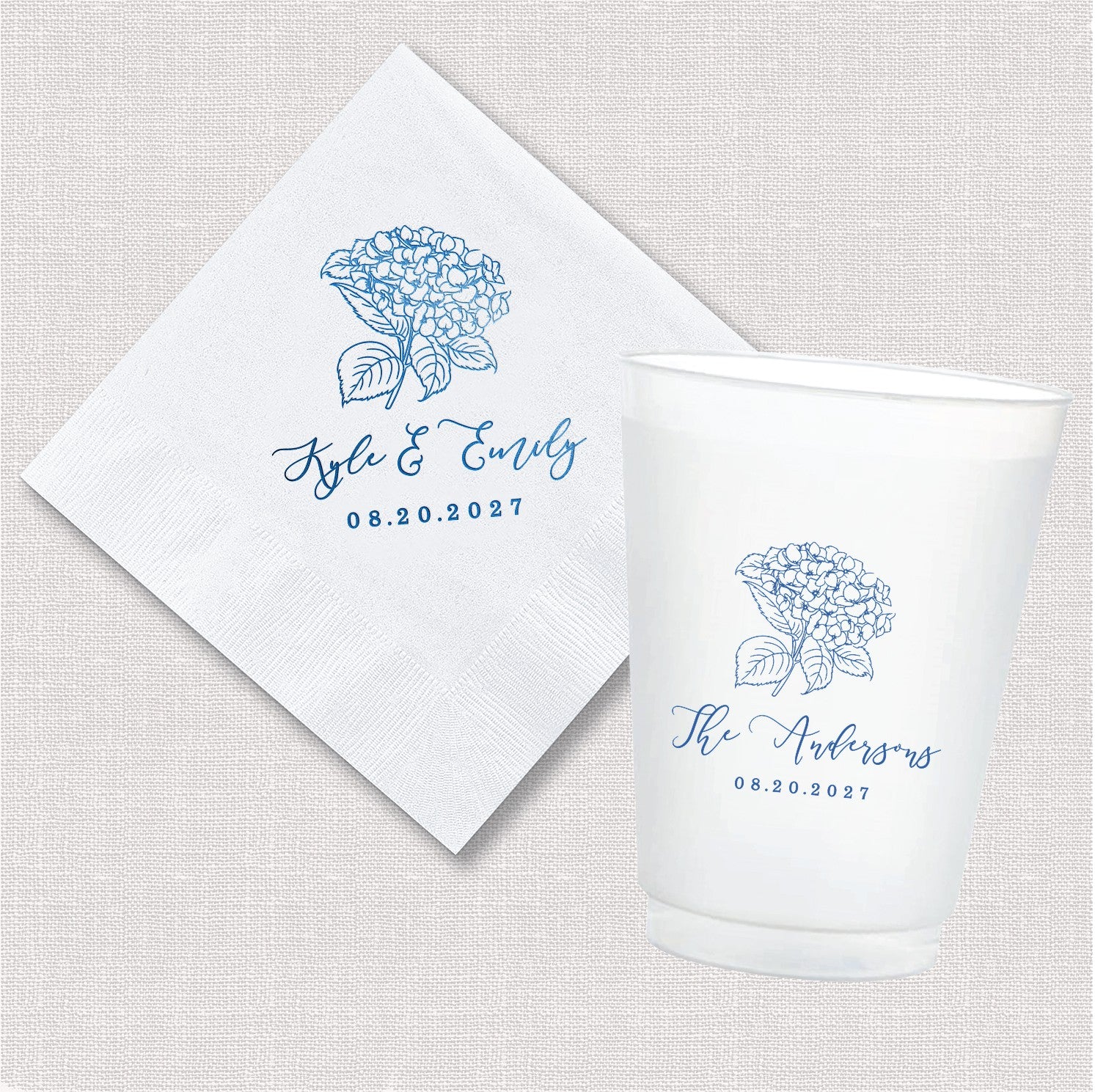 A white paper cocktail napkin and a frosted white plastic cup, both featuring a personalized blue hydrangea floral design, ideal for wedding or event decor.