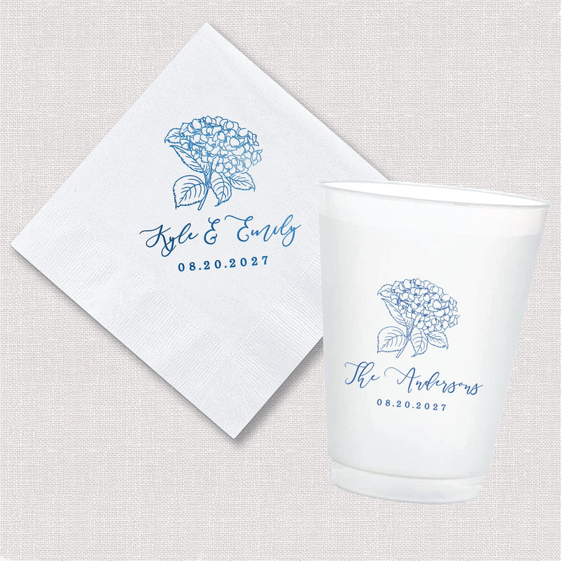 A white paper cocktail napkin and a frosted white plastic cup, both featuring a personalized blue hydrangea floral design, ideal for wedding or event decor.