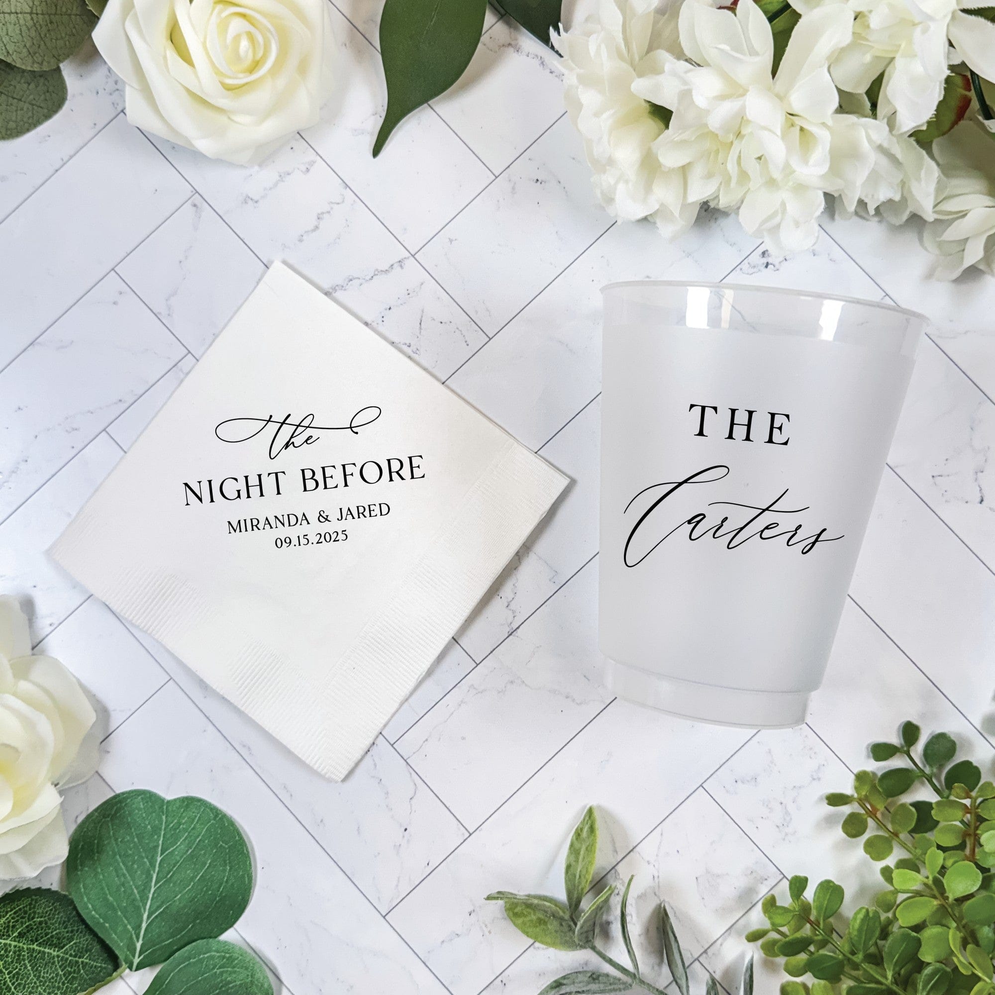 A white personalized paper napkin and a frosted white plastic cup, both featuring custom black script, are arranged on a white marble background with elegant white flowers and green foliage, ideal for a pre-wedding celebration.