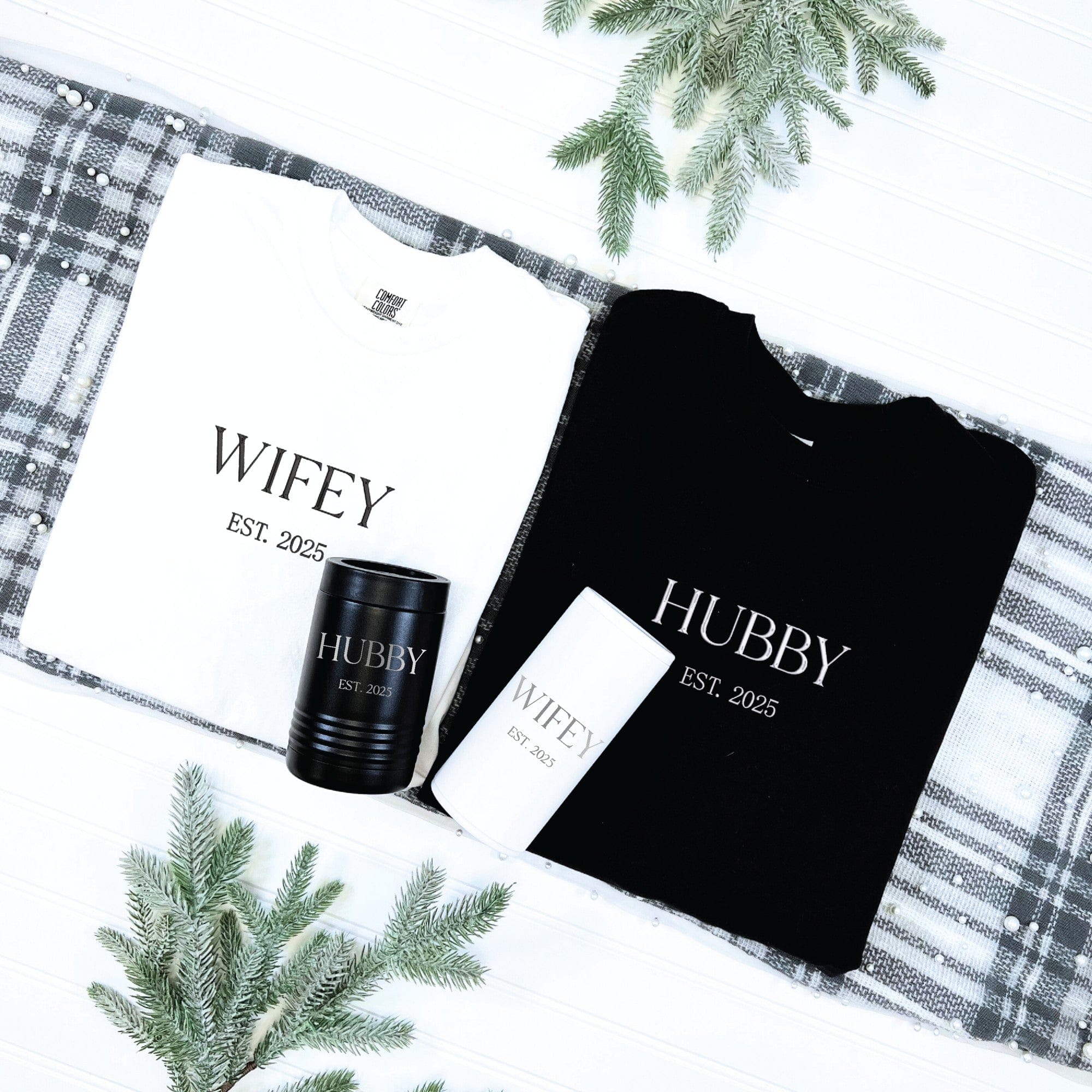 A personalized newlywed gift set including one white t-shirt, one black t-shirt, one black insulated can cooler, and one white insulated can cooler, arranged on a plaid fabric.