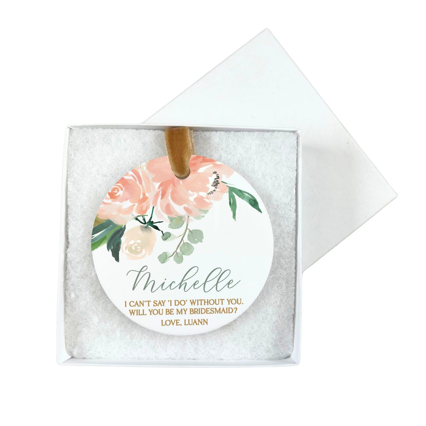 A round white ceramic ornament with a watercolor-style pink floral and green foliage design, featuring a gold velvet ribbon, presented in a white gift box with shredded paper, intended as a bridal party proposal gift.