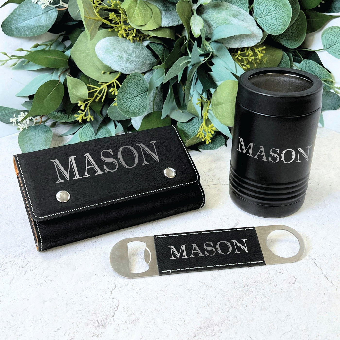 A personalized black faux leather poker chip case, a matching insulated can cooler, and a stainless steel bottle opener with faux leather detail are displayed, designed as a coordinated gift set for events.