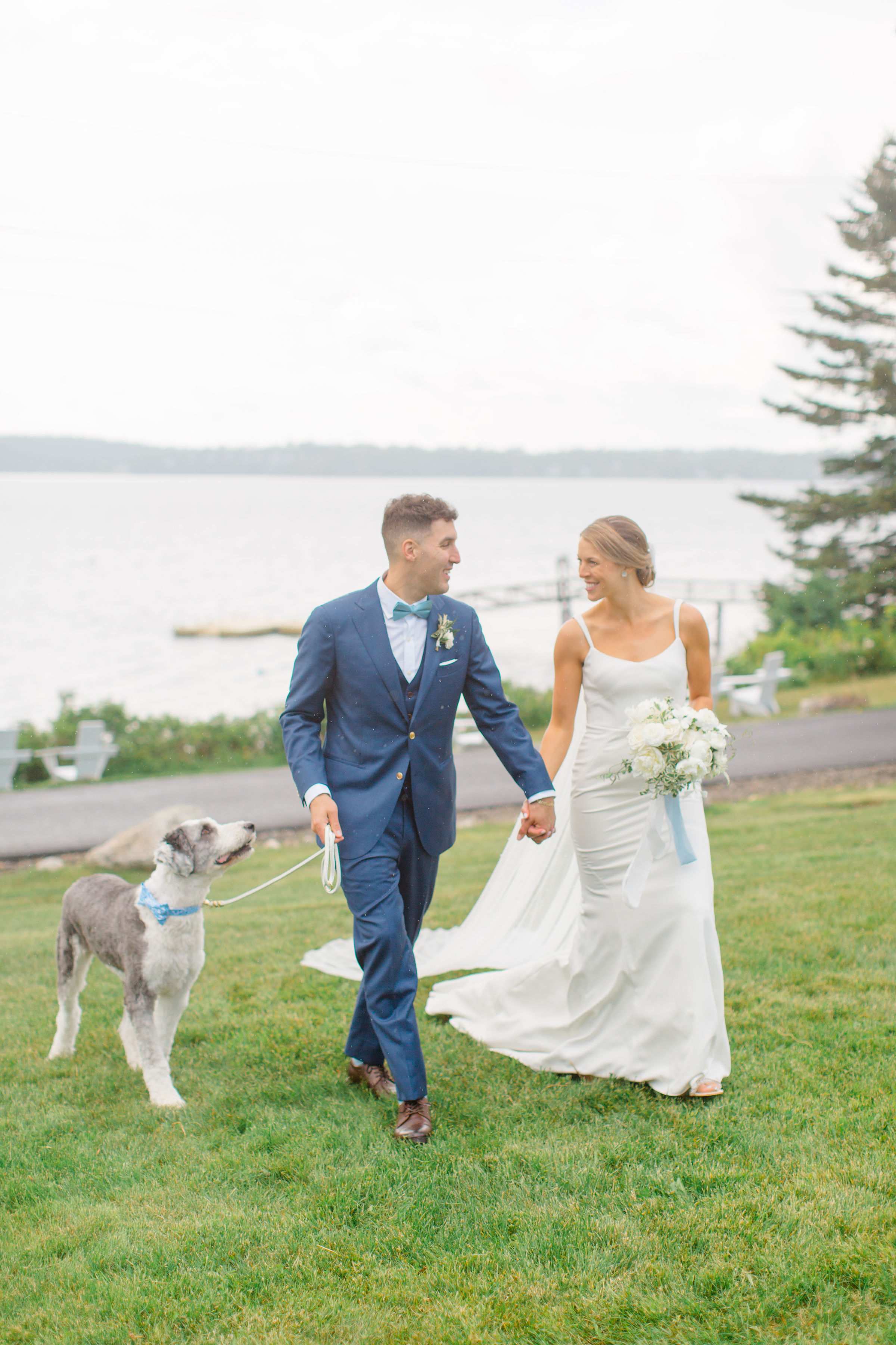 A joyful bride in a white wedding gown and groom in a navy suit walk hand in hand with their shaggy gray and white dog on a grassy lawn beside a lake during their outdoor wedding celebration.
