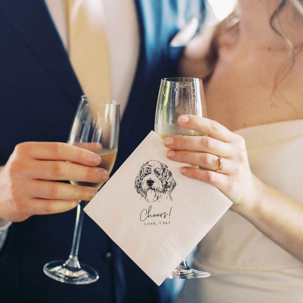 A couple holds champagne flutes, showcasing a custom dog illustration paper napkin with text, perfect for a wedding or engagement celebration.