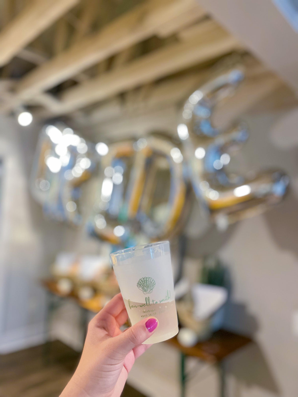 A hand holding a custom frosted plastic cup with a green seashell design and event-themed text, set against a blurred background of silver foil balloons spelling 'BRIDE' for a bridal or bachelorette party celebration.
