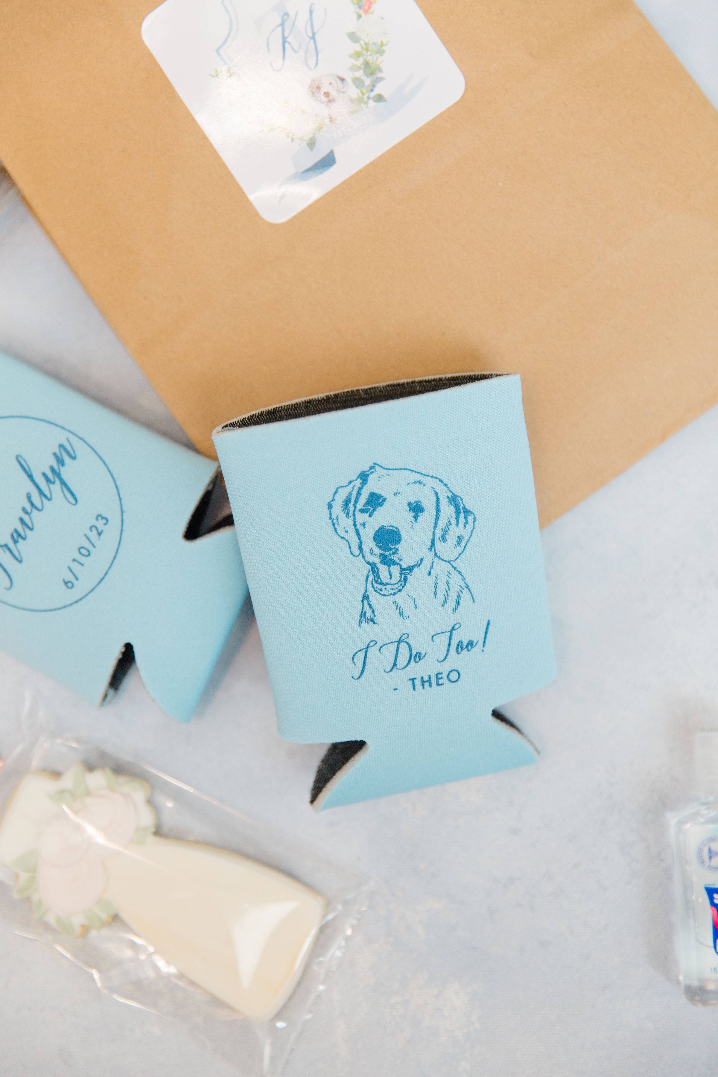 Two light blue neoprene can coolers, one featuring a custom dog illustration and special text, the other with a personalized inscription, displayed with a wedding dress-shaped cookie, ideal for wedding or engagement favors.