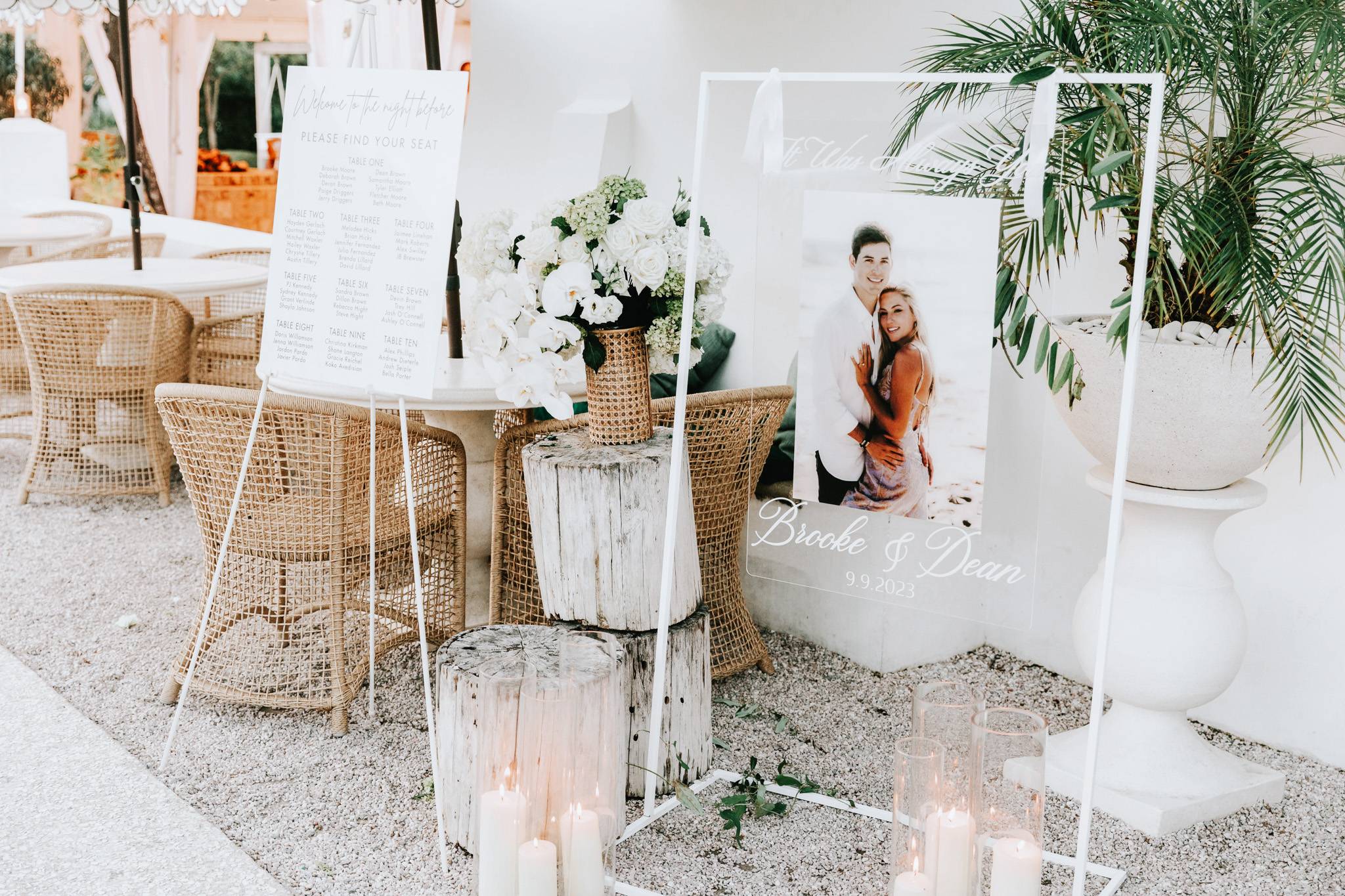 An elegant outdoor wedding reception setup showcasing a white acrylic seating chart on an easel and a clear acrylic welcome sign with a couple's photo, surrounded by wicker chairs, white floral arrangements, and candles.