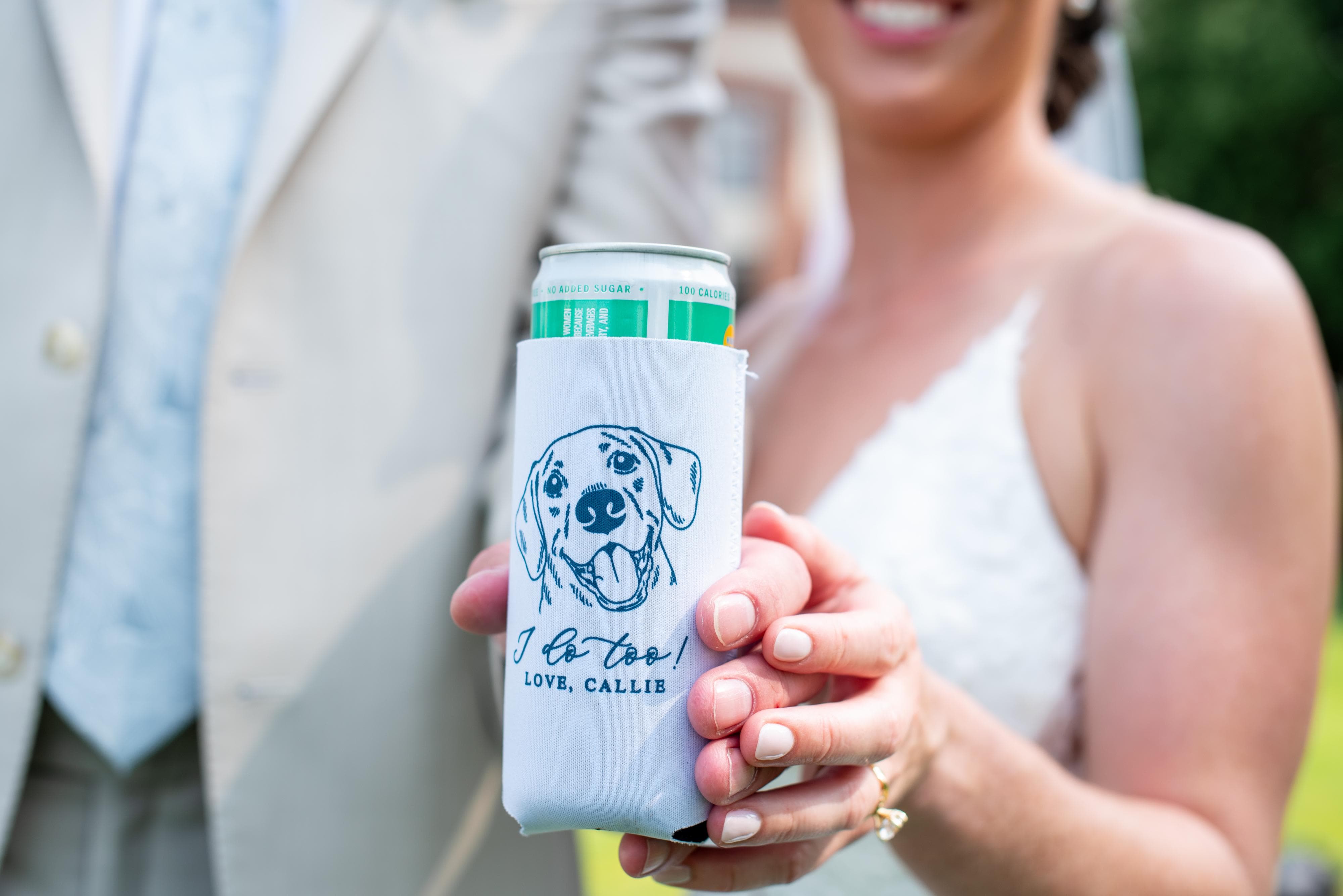 A close-up of a bride holding a personalized white foam can cooler featuring a custom dog illustration and 'I do too!' text, with a blurred groom in a suit in the background, ideal for a wedding event.