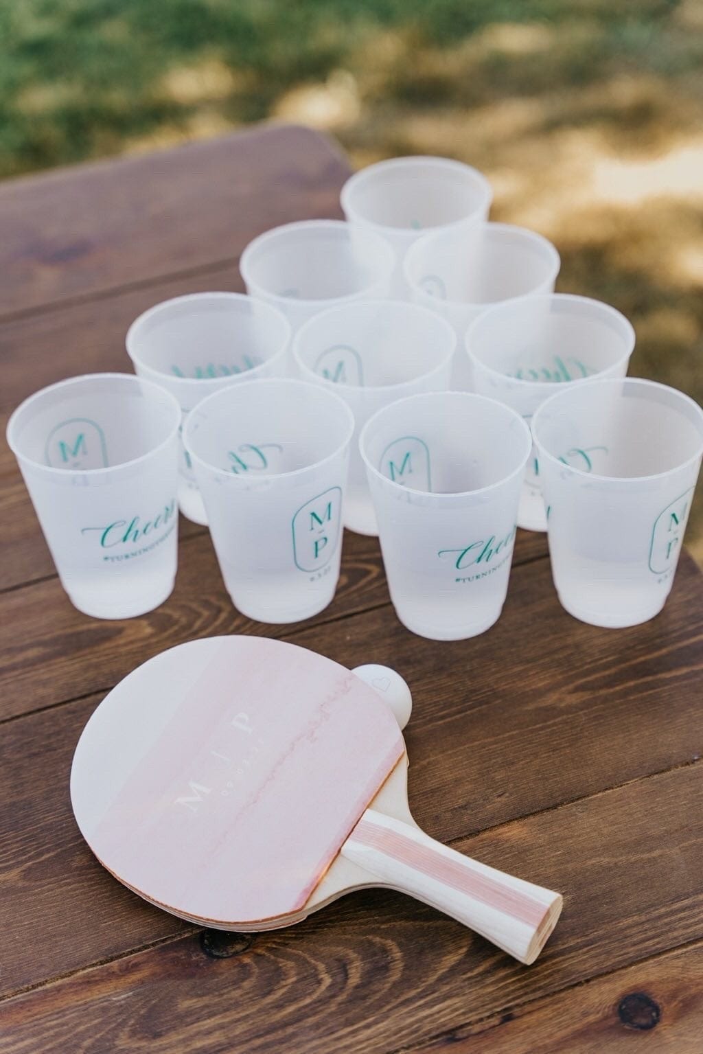 A custom-designed ping-pong paddle and frosted plastic cups with personalized teal monograms are arranged on a wooden table for an outdoor event.