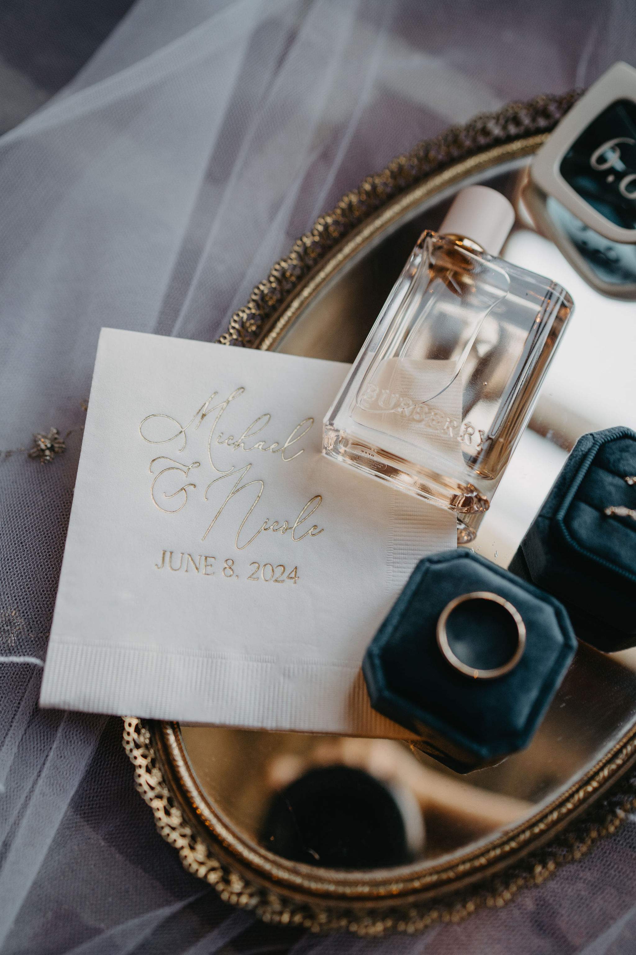 A luxurious flat lay showcasing a white personalized wedding napkin with elegant gold foil lettering, accompanied by a gold wedding band in a deep teal velvet ring box, a designer perfume bottle, and an ornate gold tray, all set on sheer white bridal fabric.
