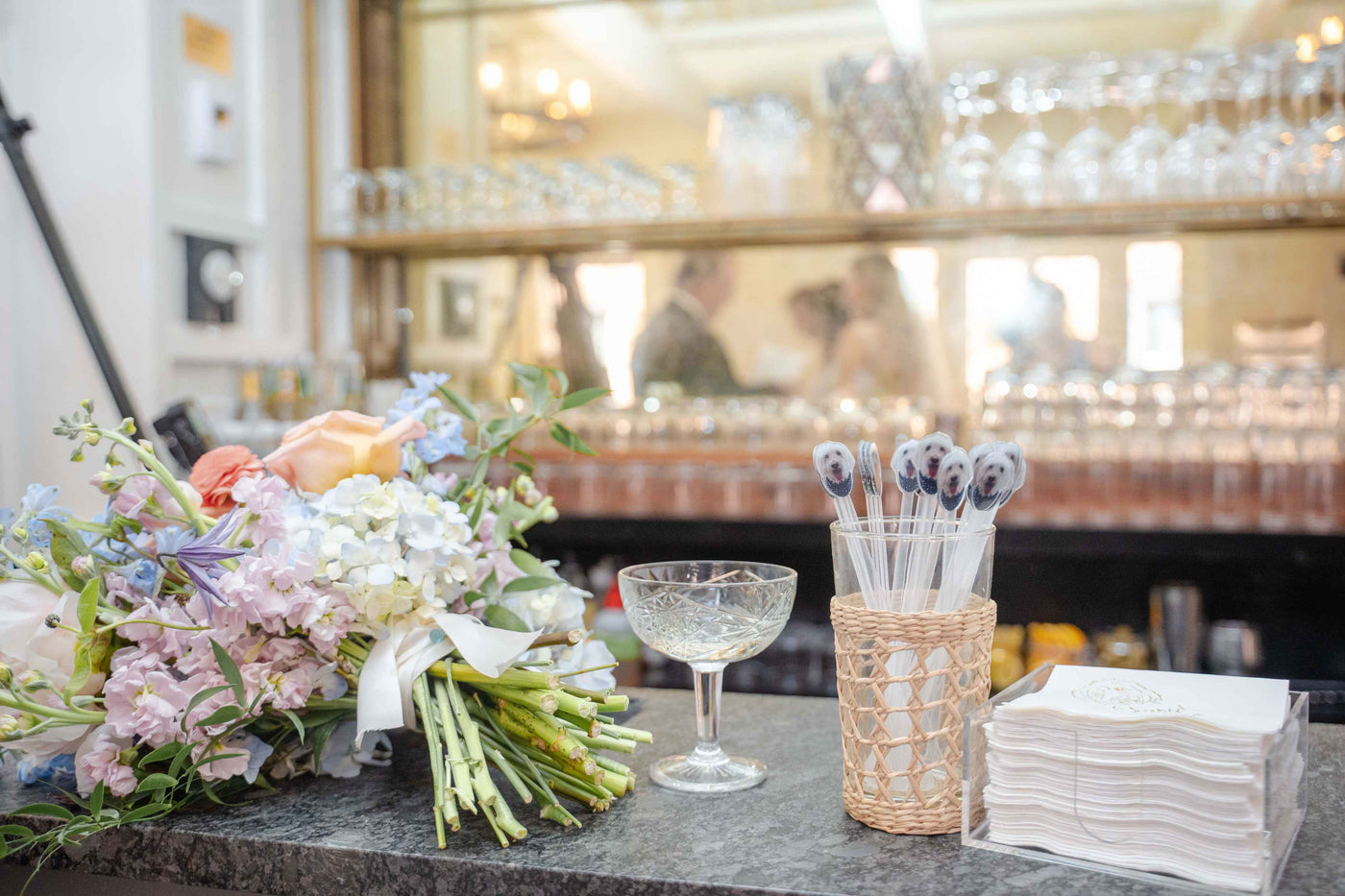 A sophisticated wedding bar setup featuring a vibrant floral bouquet, an elegant cocktail glass, personalized drink stirrers with a white dog illustration, and custom printed paper cocktail napkins, creating a festive atmosphere for guests.