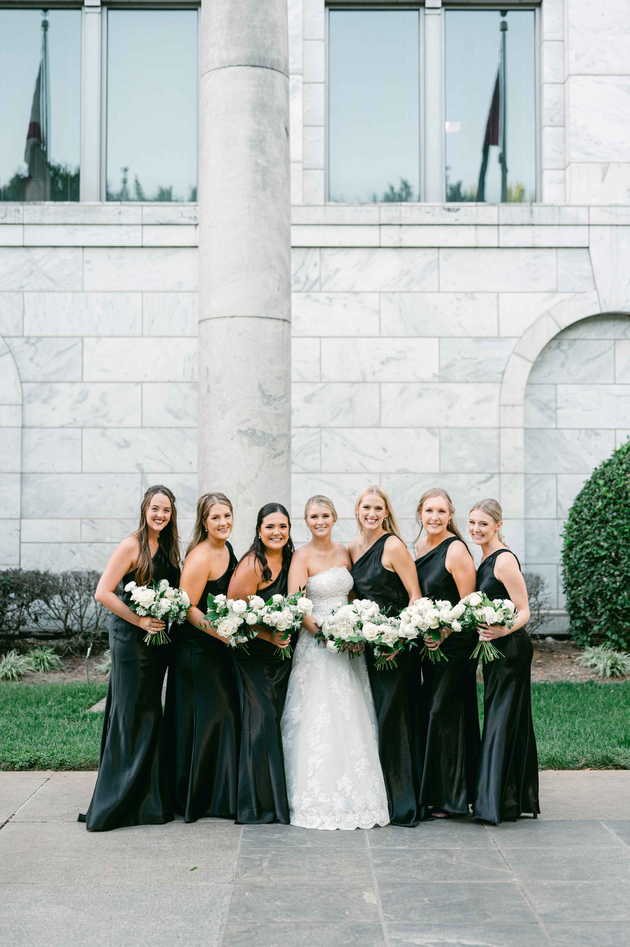 A happy bride in a white lace gown stands smiling with six bridesmaids elegantly dressed in long black dresses, each holding a white floral bouquet, against a grand white marble building backdrop at their outdoor wedding celebration.