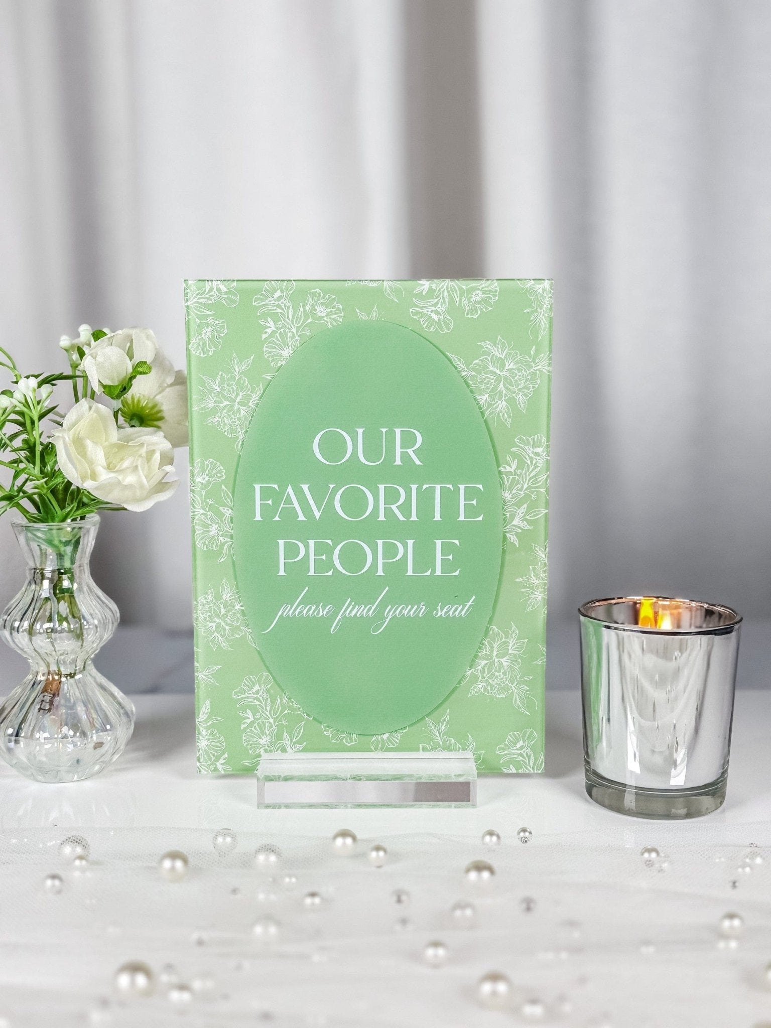 A light green acrylic wedding seating sign featuring white floral line art, a central oval design, and a clear acrylic stand.