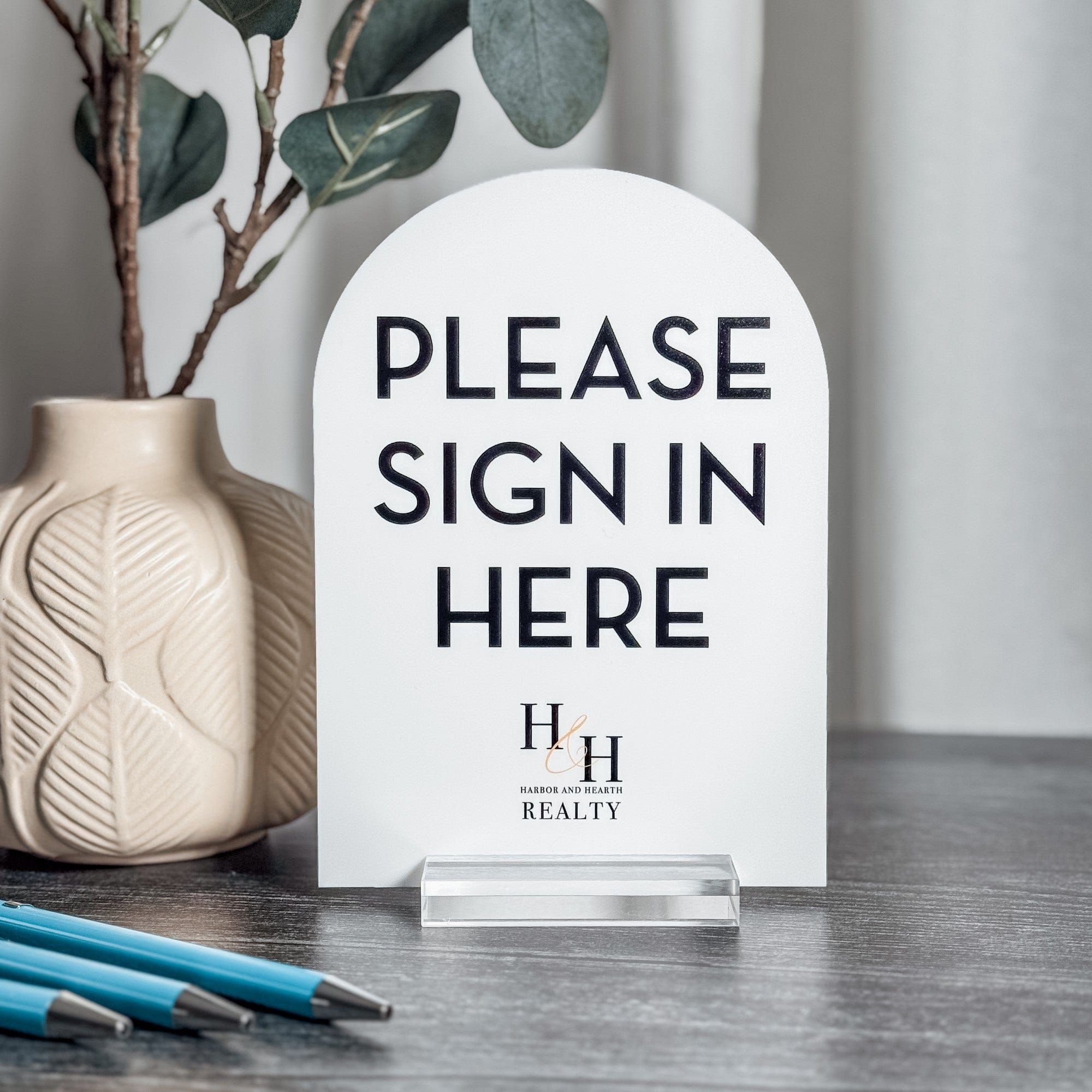 A personalized white arched acrylic sign with a clear acrylic stand, designed to indicate a guest sign-in area at a wedding or event.