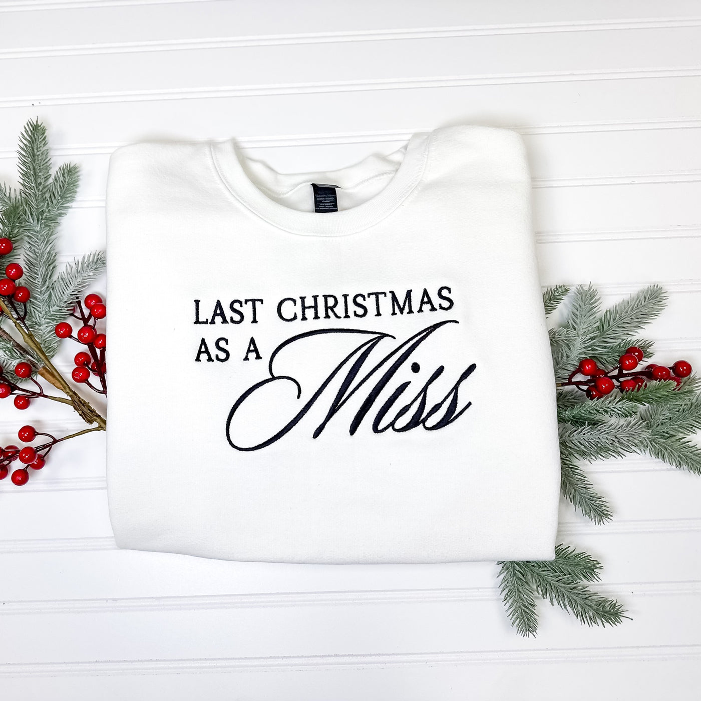 A cozy white crewneck sweatshirt featuring elegant black embroidery, designed for a bridal celebration during the Christmas season and adorned with faux holiday foliage.