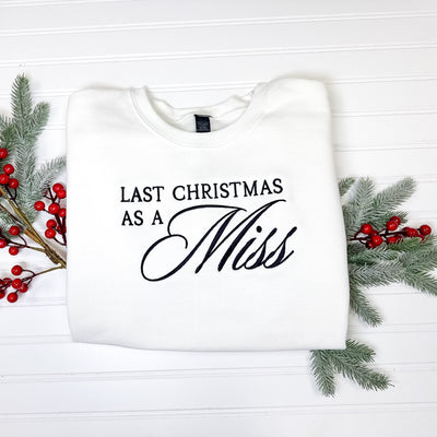 A cozy white crewneck sweatshirt featuring elegant black embroidery, designed for a bridal celebration during the Christmas season and adorned with faux holiday foliage.