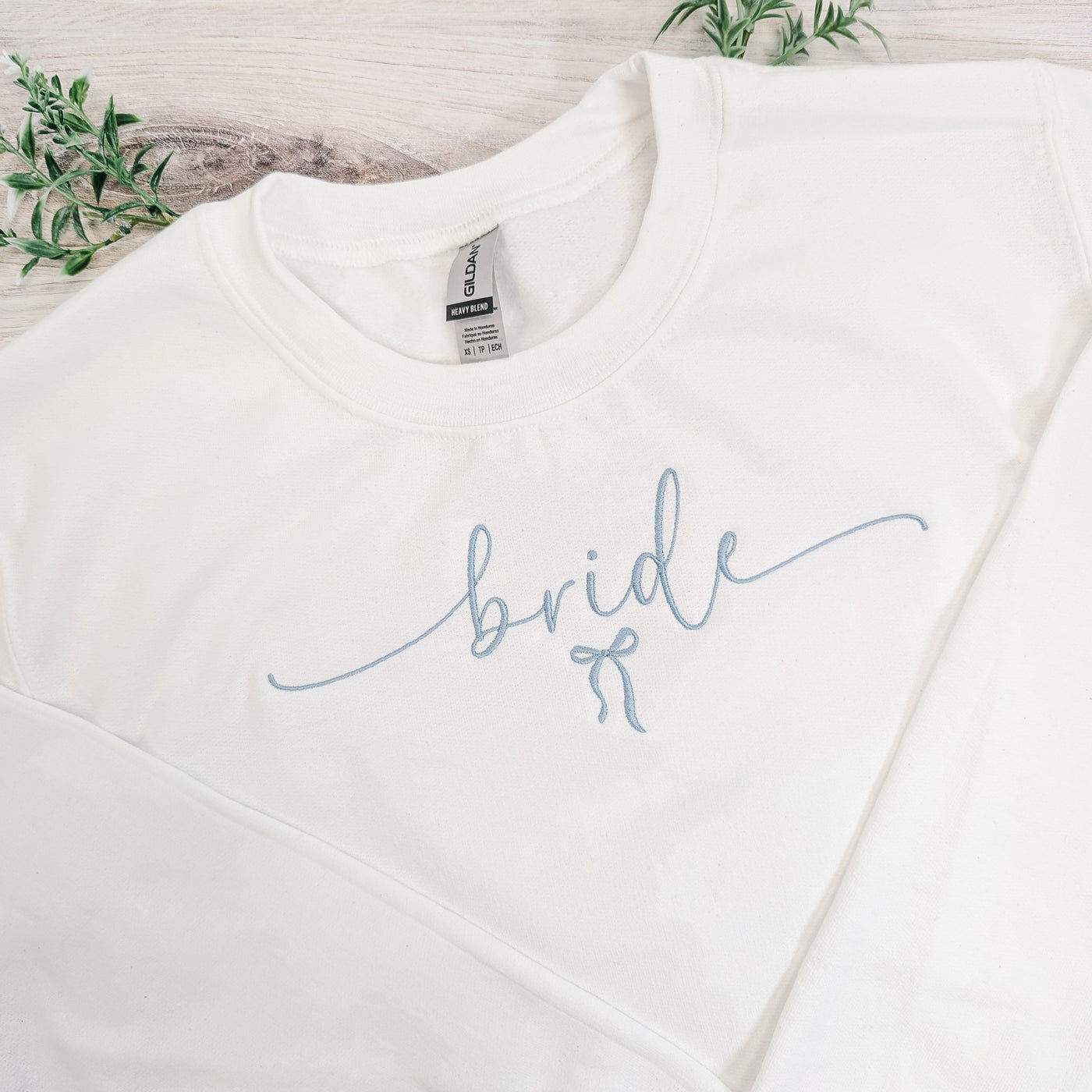 A white crewneck sweatshirt features elegant light blue script embroidery reading 'bride' with a small bow design, perfect for a wedding or bridal shower.
