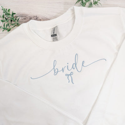 A white crewneck sweatshirt features elegant light blue script embroidery reading 'bride' with a small bow design, perfect for a wedding or bridal shower.