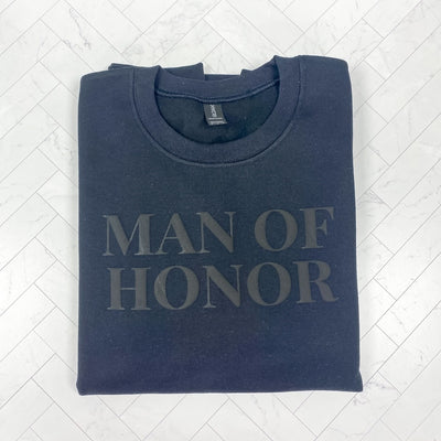 A folded black crewneck sweatshirt made of a soft cotton blend, featuring a subtle, tone-on-tone black design ideal for a member of a wedding party.