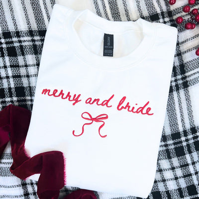 A white crewneck sweatshirt featuring red embroidered lettering and a bow design, suitable bridal apparel for a festive wedding occasion.