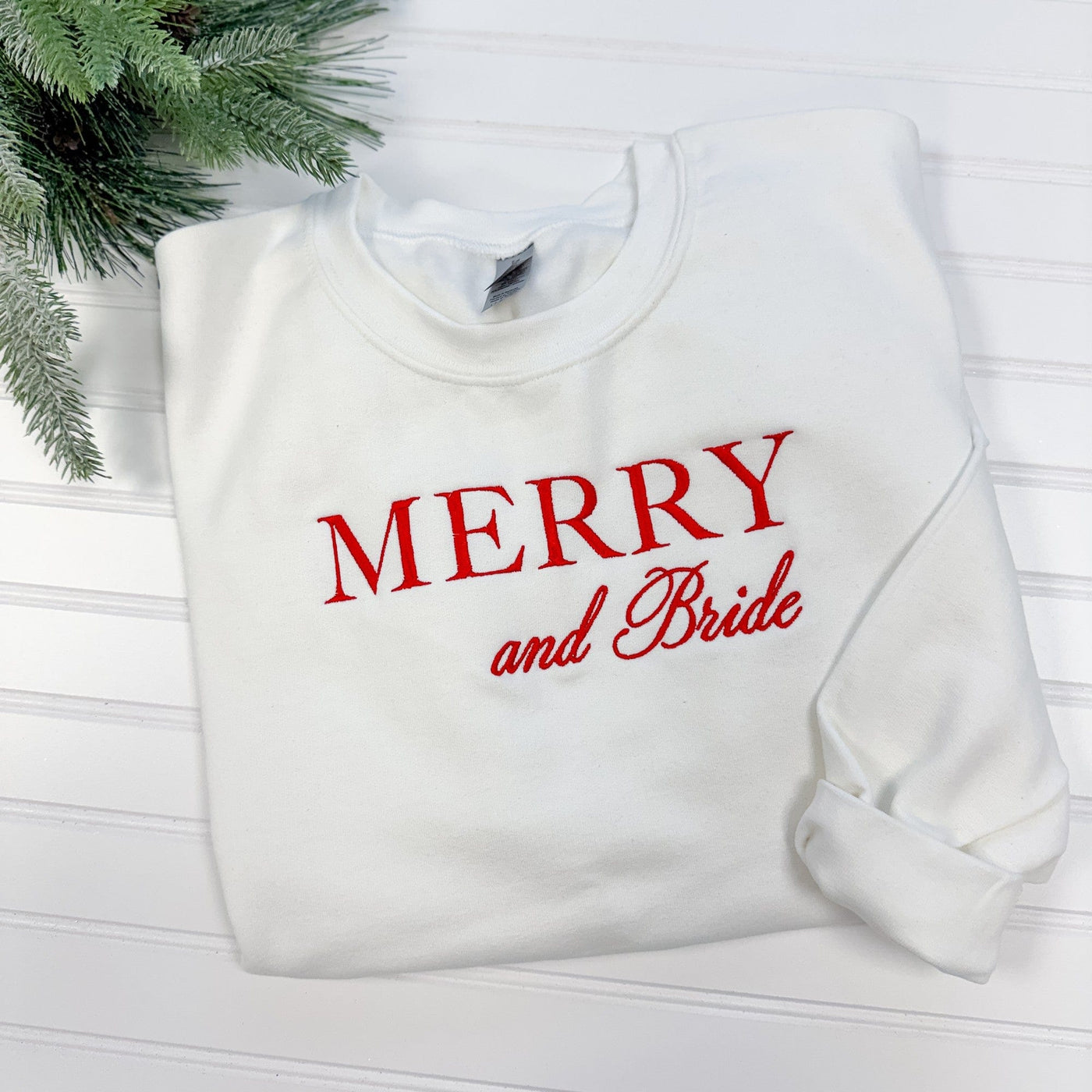 A white crewneck sweatshirt made from soft fabric, featuring festive red embroidered lettering, ideal for bridal wear during a winter celebration.