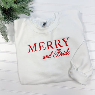 A white crewneck sweatshirt made from soft fabric, featuring festive red embroidered lettering, ideal for bridal wear during a winter celebration.