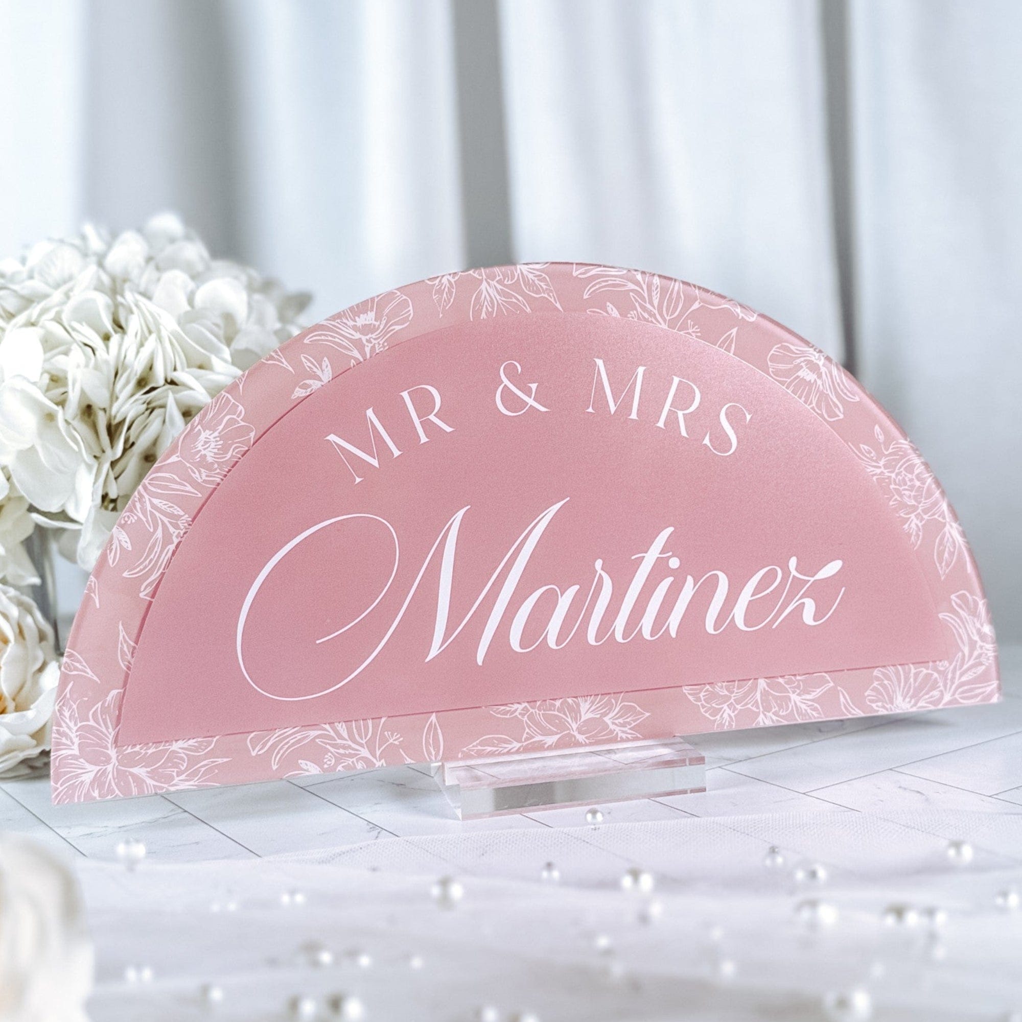 A blush pink semi-circular acrylic wedding sign featuring elegant white floral outlines and personalized details, displayed on a clear acrylic stand.