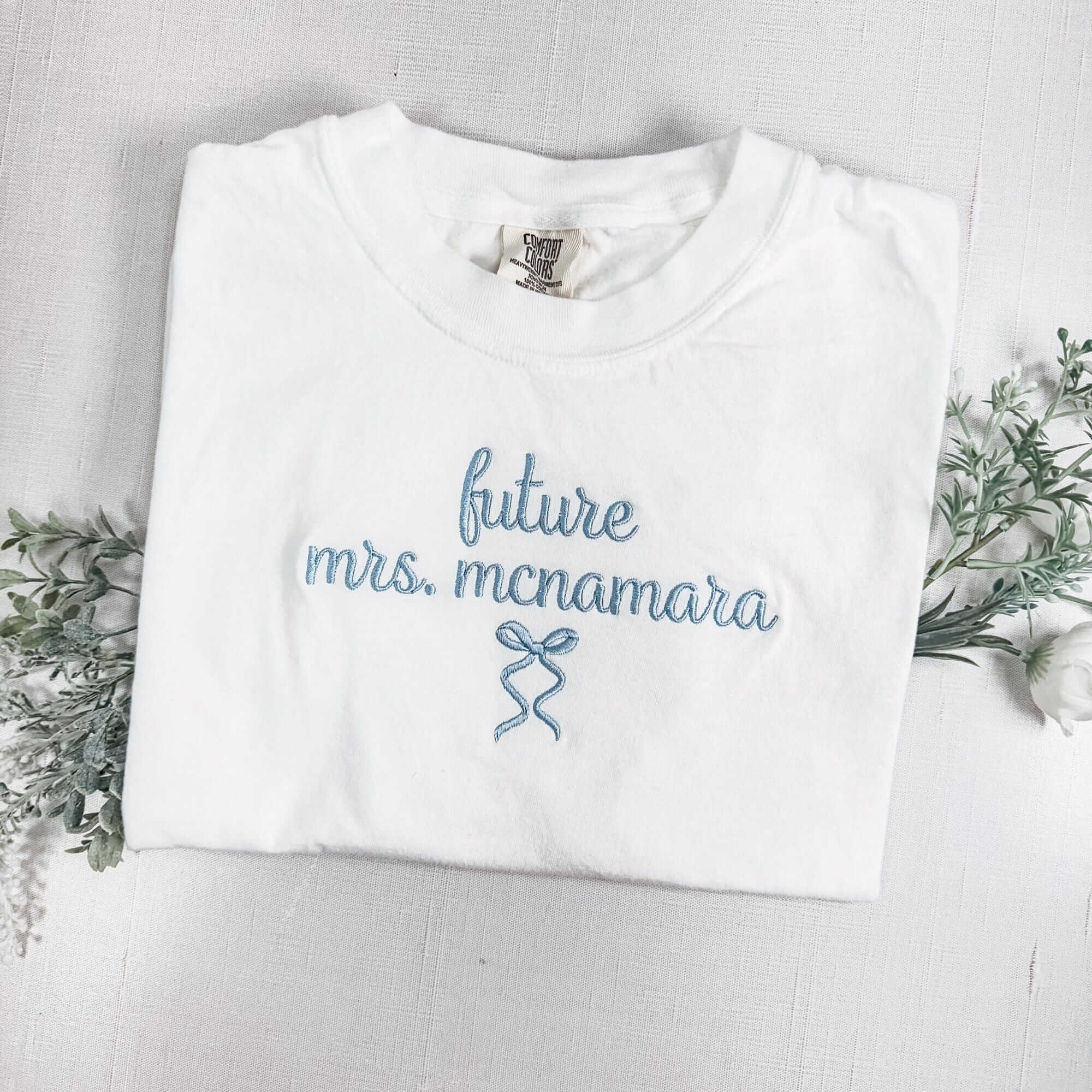 A folded white cotton t-shirt featuring light blue personalized embroidery and a decorative bow design, ideal for a bridal event.