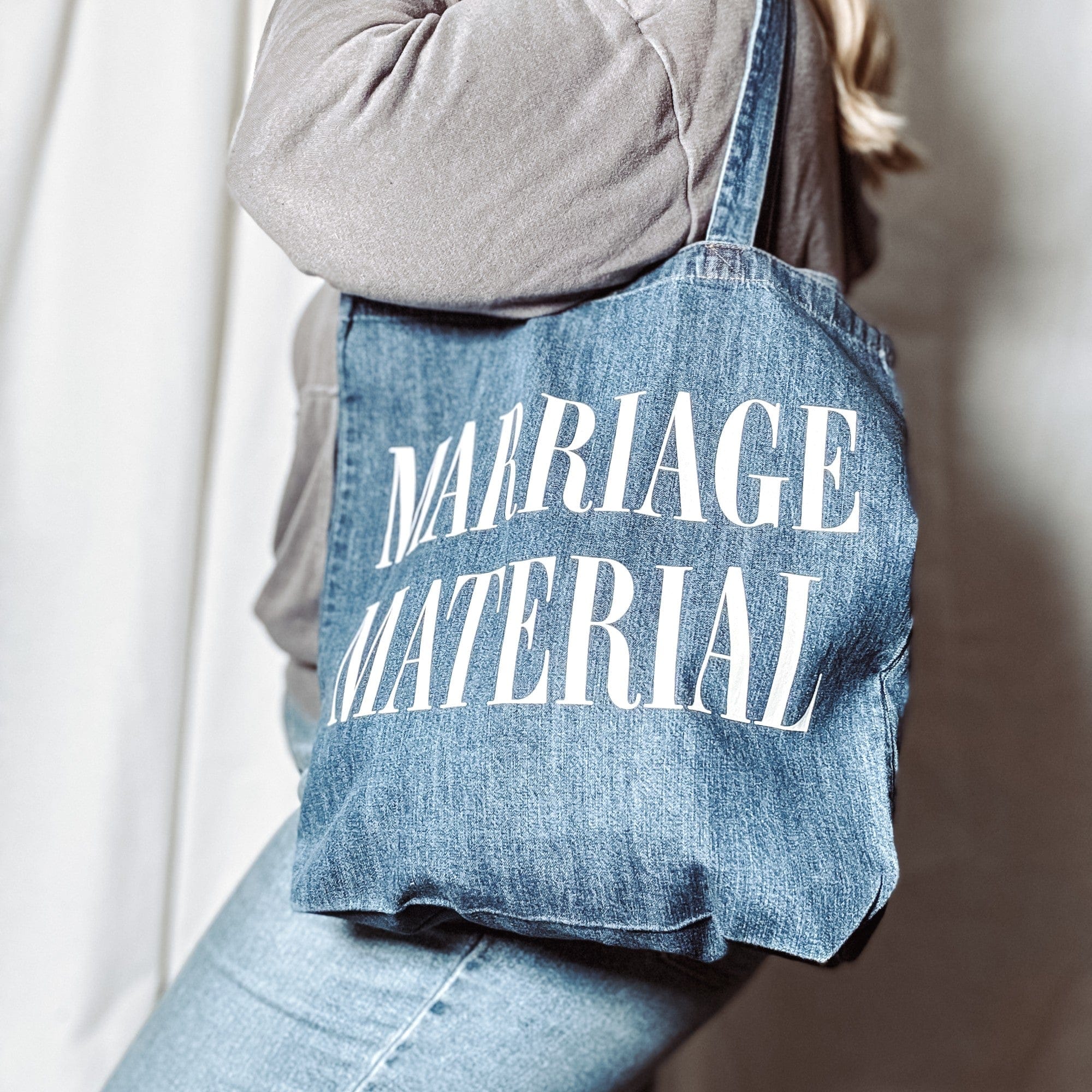 A person wears a casual denim-style tote bag with shoulder straps, featuring a personalized white design, perfect as a functional accessory for a wedding or bridal event.