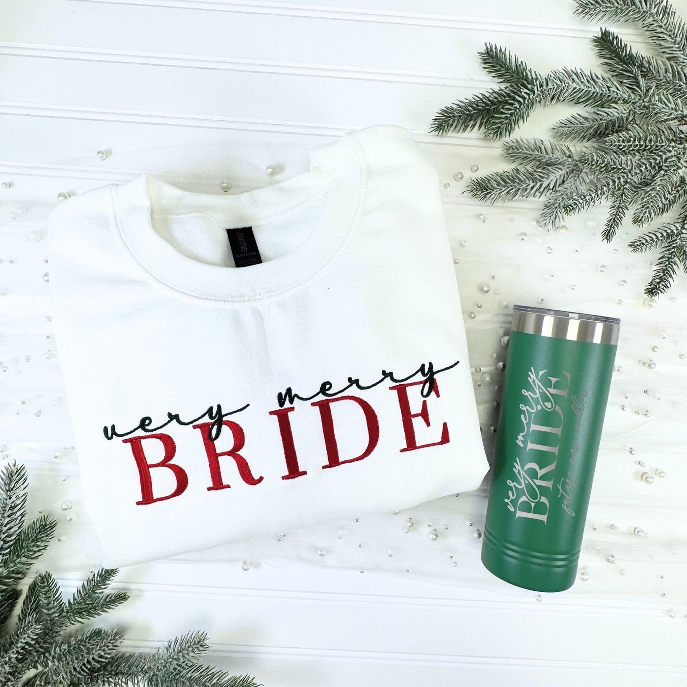A festive bridal gift set featuring a white long-sleeve sweatshirt and a dark green insulated tumbler, both customized for a winter-themed wedding, displayed on a white background with frosted evergreen branches and pearls.