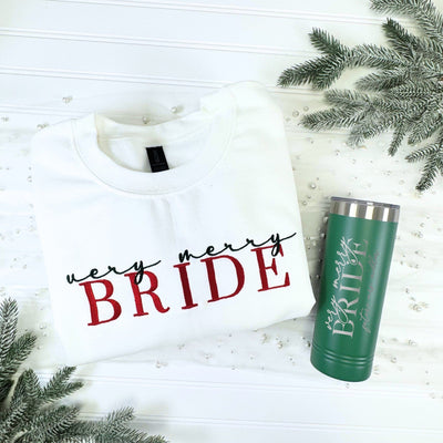 A festive bridal gift set featuring a white long-sleeve sweatshirt and a dark green insulated tumbler, both customized for a winter-themed wedding, displayed on a white background with frosted evergreen branches and pearls.