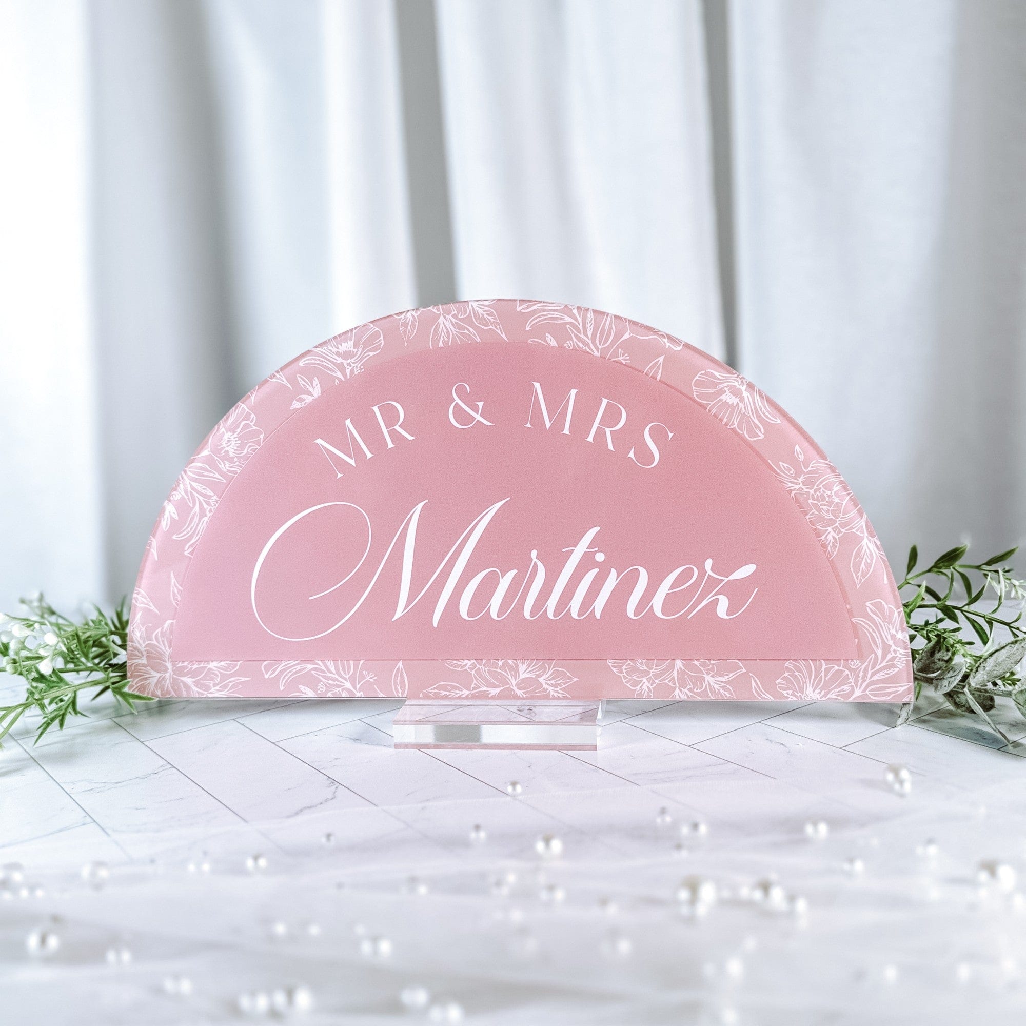 A semicircular pink acrylic wedding sign featuring a white floral line art border and white script text, displayed on a clear stand amidst greenery and pearls.