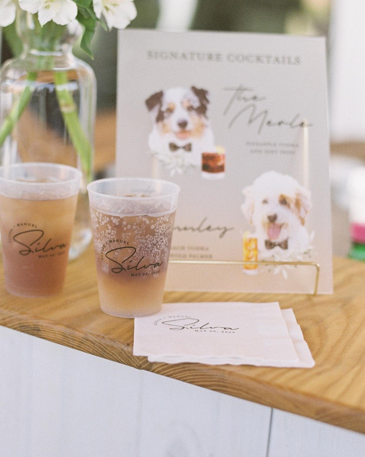 A personalized wedding bar set featuring custom frosted plastic cups, white paper napkins, and a signature cocktail menu sign with charming pet illustrations, designed to add a unique and cohesive touch to event beverages.