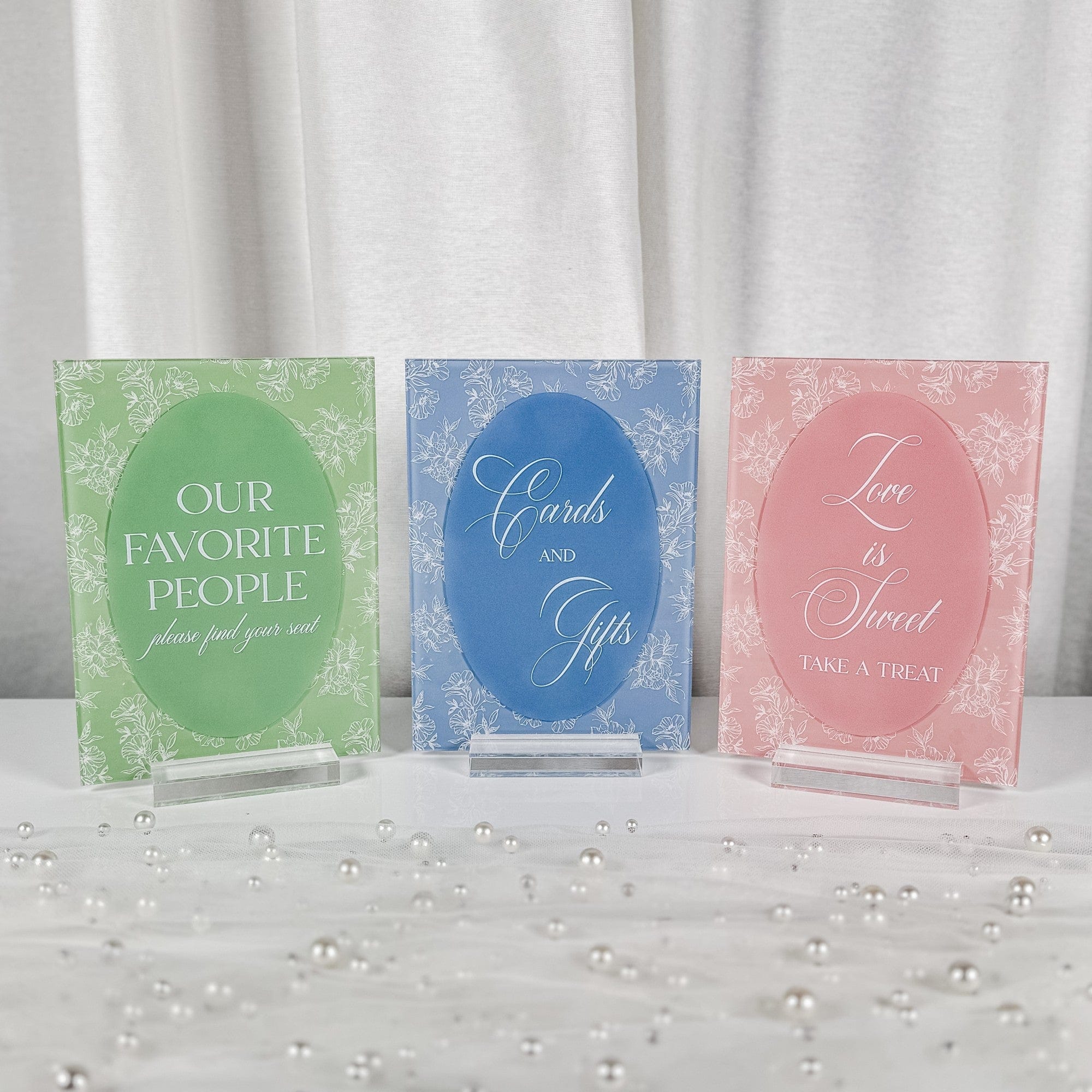 A set of three rectangular acrylic wedding decor signs in pastel green, blue, and pink, each featuring a white floral pattern and a central oval design, displayed on clear acrylic bases.
