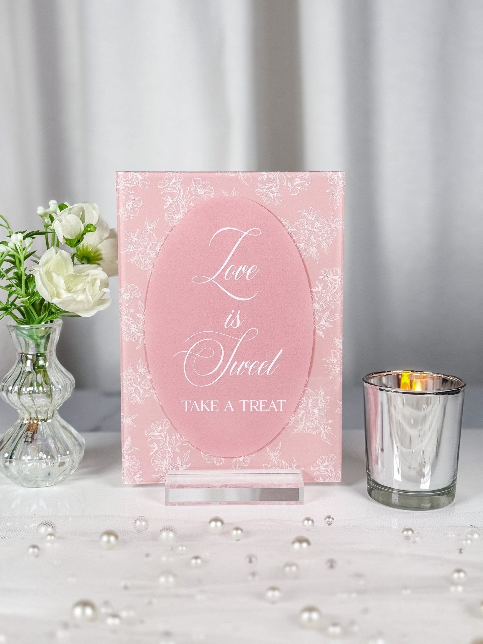 A vertical rectangular pink acrylic sign with a white floral line art background and a contrasting solid pink oval overlay, presented on a clear acrylic stand for a wedding or event dessert table.