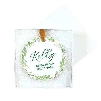 A circular white tag featuring a watercolor green foliage and light pink floral wreath design, attached to a brown velvet ribbon, serving as a personalized wedding or bridal shower favor or gift tag.