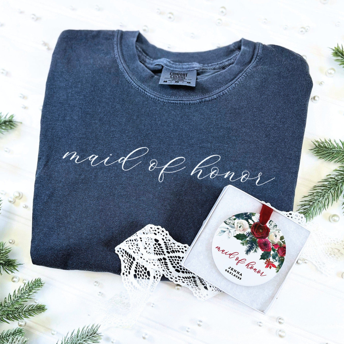 A comfortable dark gray crewneck sweatshirt features a decorative white script design on the front, accompanied by a gift box containing a round white ornament adorned with a red and white floral pattern and a red velvet ribbon, forming a complete wedding party gift set.