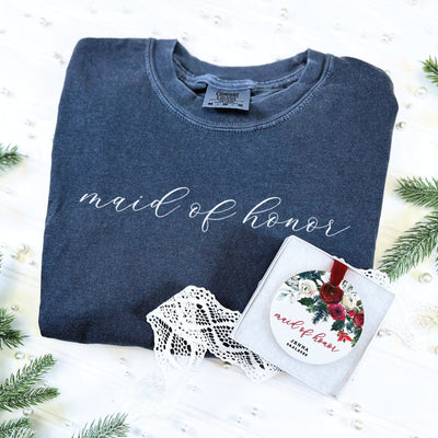 A comfortable dark gray crewneck sweatshirt features a decorative white script design on the front, accompanied by a gift box containing a round white ornament adorned with a red and white floral pattern and a red velvet ribbon, forming a complete wedding party gift set.