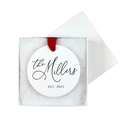 A round white personalized ornament with a black script design and a red velvet ribbon is presented in a white gift box with soft filler, ideal for a wedding or special occasion keepsake.