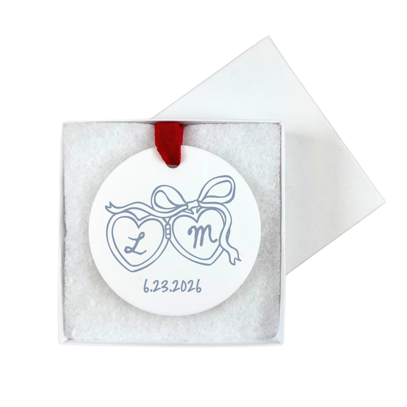 A white round ceramic wedding ornament featuring two hearts connected by a bow design, hangs by a red ribbon and is presented in a white gift box with shred filling.