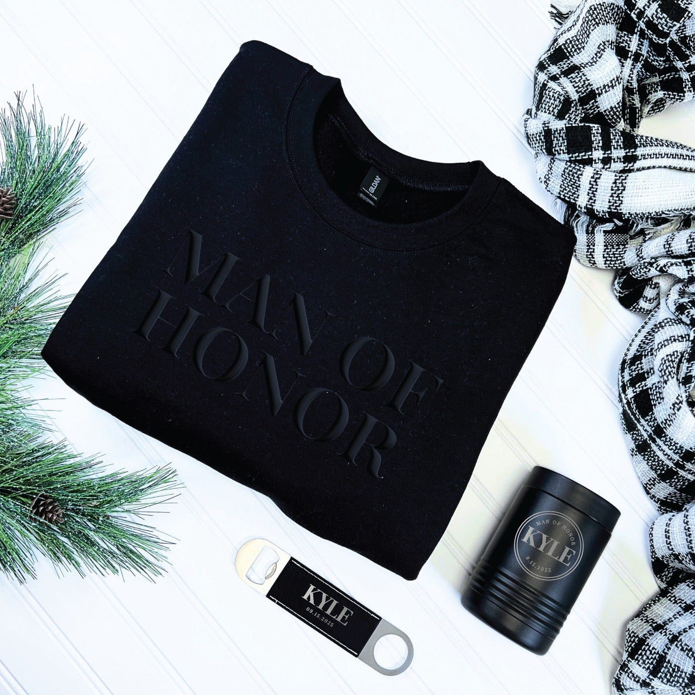 A black long-sleeved sweatshirt with a raised text design is displayed alongside a black insulated can cooler with an engraved design and a silver bottle opener with a black leatherette grip, all part of a personalized wedding party gift set.